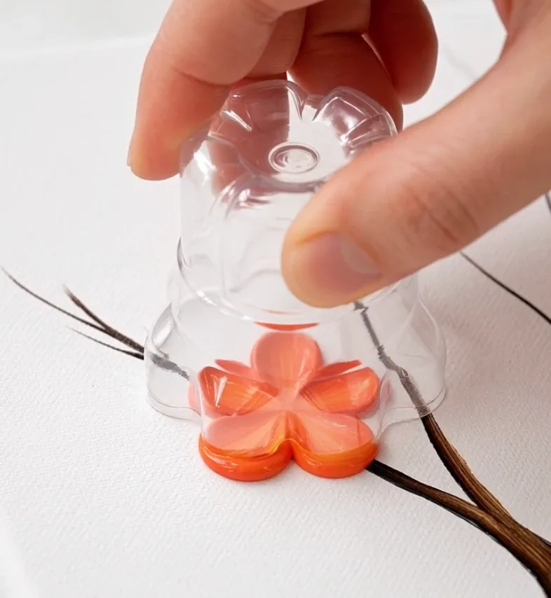 Artist stamping five-petal blossoms onto a painted tree using a plastic bottle base